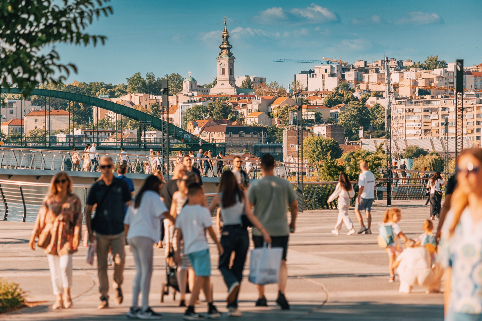 Gente paseando por el paseo del Sava en Belgrado, con el casco antiguo y la torre de una iglesia al fondo